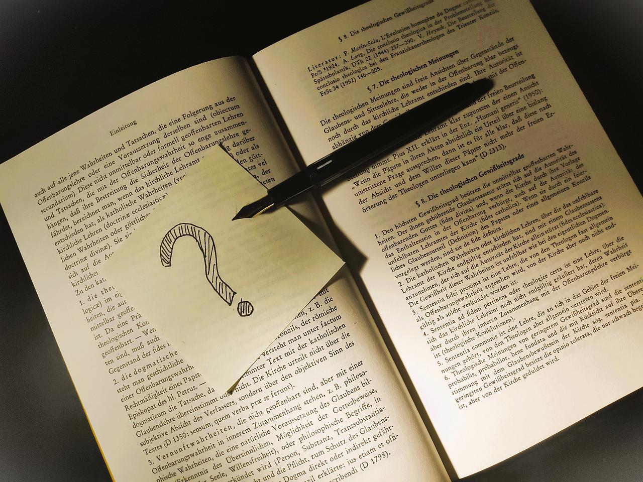Ein Zettel mit gezeichnetem Fragezeichen und Füllfederhalter auf einem augeschlagenen Buch.