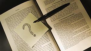 Ein Zettel mit einem gemalten Fragezeichen und ein Füllfederhalter auf einem aufgeschlagenen Buch.