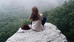 Frau sitzt auf einem Felsen von einem Wald, neben ihr ein roter Hut.
